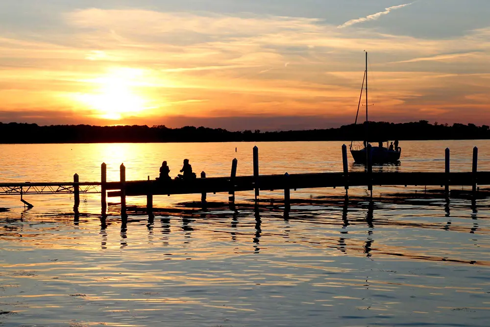 silhouette of couple at end of boat dock with sailing boat in background at sunset. With water, obviously.